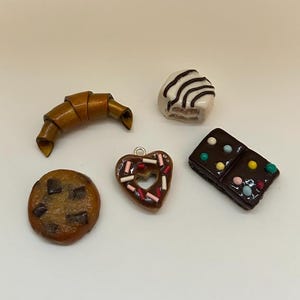 May include: A collection of miniature food charms, including a croissant, a chocolate chip cookie, a heart-shaped pastry with sprinkles, a brownie, and a small cake with chocolate stripes. The charms are in various shades of brown and white.