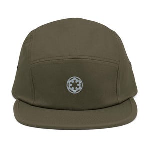 May include: An olive green five-panel cap featuring a flat brim and a centered, light gray embroidered circular emblem depicting a stylized six-spoked gear design. The hat is constructed from a solid-colored fabric with visible stitching details.