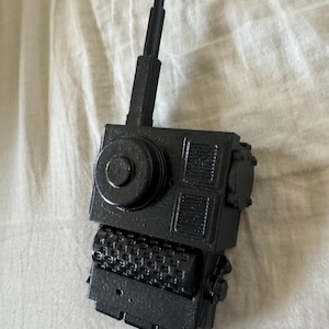 May include: A matte black handheld communication device featuring a prominent vertical antenna, a large circular dial, two rectangular speaker grilles, and a textured cylindrical roller component positioned at the base of the main rectangular housing.
