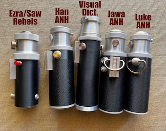 May include: Five cylindrical black and silver handheld devices with metallic top sections and various side-mounted knobs, buttons, and rings. Text labels above the items read Ezra/Saw Rebels, Han ANH, Visual Dict., Jawa ANH, and Luke ANH.