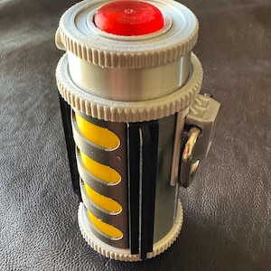 May include: A cylindrical prop featuring a bright red circular top button, a silver-toned body with vertical black accents, and a central section displaying four yellow oval cutouts. A small silver metal ring is attached to the side.