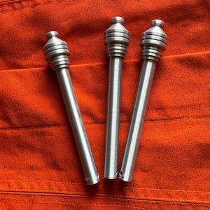 May include: Three silver-colored metal objects with a cylindrical design and a tiered, rounded top. The objects are arranged on an orange fabric surface. The objects have a long, slender body with a small hole at the bottom.