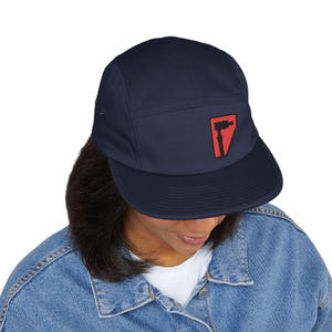 Peut inclure: Une casquette bleu marine à cinq panneaux avec une calotte structurée et une visière plate. Au centre du panneau avant se trouve un écusson triangulaire à fond rouge arborant une silhouette noire représentant un outil, tel qu'une perceuse.