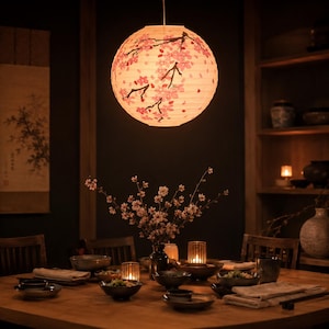 Könnte beinhalten: Eine runde Papierlaterne mit Kirschblütendesign hängt über einem Holzesstisch. Die Laterne ist von innen beleuchtet und wirft ein warmes Licht. Der Tisch ist mit Schalen, Kerzen und einer Blumenvase gedeckt, was eine gemütliche Atmosphäre schafft.