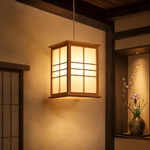 May include: A square, wooden pendant light fixture with a grid pattern. The light has a warm, inviting glow, and is suspended from the ceiling by a thin cord. The fixture is a traditional Japanese design, and is set against a neutral-toned wall.