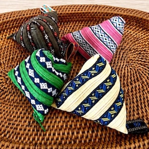 Puede incluir: Cuatro bolsas triangulares con distintos patrones de tela y colores. Una es gris oscuro con bordados florales rojos y blancos, una rosa con bandas geométricas, una azul marino con ribete verde y motivos blancos, y una crema con rayas azules.