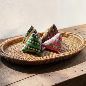 May include: Four small, triangular-shaped fabric pouches with zippers, featuring colorful woven patterns in green, pink, brown, and blue, arranged on a large, oval-shaped, light brown woven rattan serving tray with two side handles.