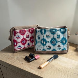 May include: Two rectangular fabric pouches with beige sides and patterned front panels. One panel features a red and white print with icons, while the other has a teal and white print with icons and the word BAO. A makeup brush and two lipsticks lie in front.