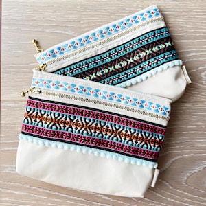 May include: Two off-white canvas zipper pouches featuring decorative woven trim. The top pouch has a blue, black, and yellow pattern with light blue pom-pom trim. The bottom pouch has a pink, orange, and black pattern with light blue pom-pom trim.