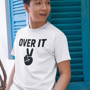 Op de afbeelding: Wit T-shirt met de tekst "OVER IT" boven een zwart vredesteken. Het shirt heeft korte mouwen en lijkt van katoen te zijn. De persoon draagt het shirt voor een blauwe luik.