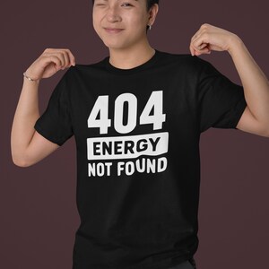 Può includere: T-shirt nero con la scritta bianca "404 ENERGY NOT FOUND". La maglietta è a girocollo e viene tenuta da una persona. Lo sfondo è color amaranto.