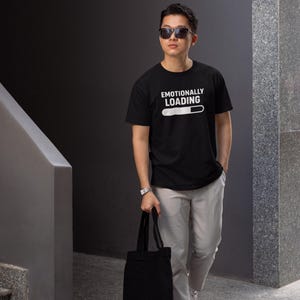Puede incluir: Camiseta negra con el texto blanco "EMOTIONALLY LOADING" y una barra de carga. La persona lleva gafas de sol, un reloj, pantalones claros y zapatillas blancas. Lleva un bolso tote negro.