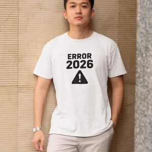 Op de afbeelding: Wit t-shirt met de tekst "ERROR 2026" boven een zwart uitroepteken. Het shirt wordt gedragen door een persoon in lichtgekleurde broek. De persoon leunt tegen een getextureerde muur.