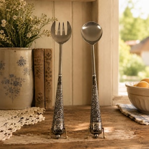 May include: A pair of silver-toned salad serving utensils featuring a three-pronged fork and a round spoon. Each utensil has a long, slender neck and a conical handle decorated with intricate, embossed floral patterns and small metal feet.
