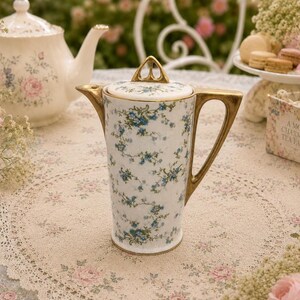 Peut inclure: Une cafetière en porcelaine haute et cylindrique ornée d'un motif délicat de petites fleurs bleues et de vignes vertes sur fond blanc. Elle possède une anse dorée, un couvercle assorti avec un bouton triangulaire et un bec verseur doré.