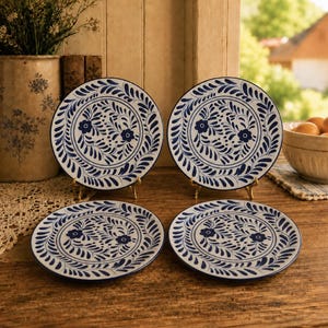 May include: A set of four round ceramic plates featuring a white base with a hand-painted dark blue floral and leaf pattern. Two plates are displayed upright on small metal stands, while two plates lie flat on a wooden surface, showcasing the design.