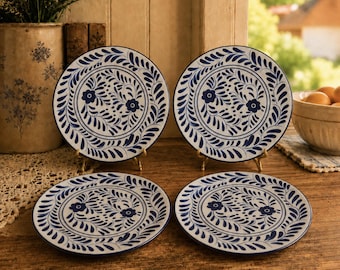 May include: A set of four round ceramic plates featuring a white base with a hand-painted dark blue floral and leaf pattern. Two plates are displayed upright on small metal stands, while two plates lie flat on a wooden surface, showcasing the design.