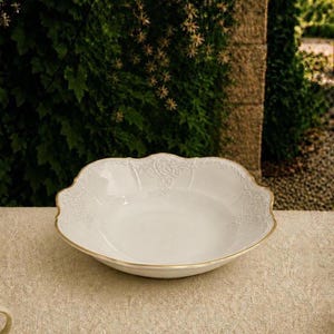 May include: A white ceramic bowl with a scalloped edge and a thin gold rim. The bowl features a subtle, embossed floral pattern along the interior rim, providing a textured detail against the smooth, glossy white surface of the basin.