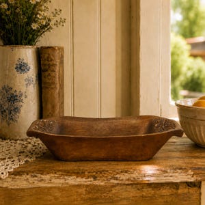 May include: A rectangular brown wooden dough bowl with carved leaf details on the handles. The bowl has a smooth, dark finish and a shallow, elongated basin. It is positioned horizontally, showcasing its natural wood grain and carved accents.