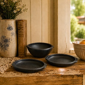 May include: A set of three dark charcoal-colored ceramic kitchen items consisting of one deep round bowl and two shallow circular plates, each featuring small side handles. The items have a matte finish and a simple, functional design.