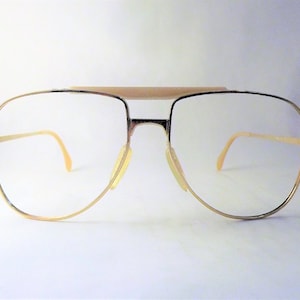 May include: Gold-rimmed spectacles with a double bridge and clear lenses. The frames have a classic aviator shape with a light-coloured brow bar and nose pads. The temples are thin and curve down to the ear. The overall design is vintage and stylish.