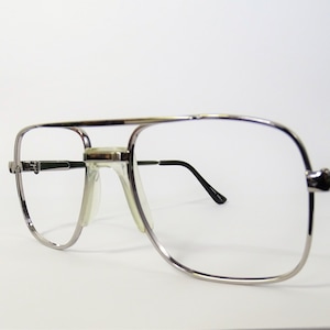 May include: Silver-framed eyeglasses with a double bridge and large, rectangular lenses. The temples are black, and the nose pads are clear. The glasses have a classic, retro style.
