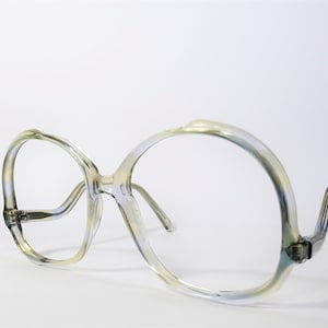 May include: Oversized eyeglasses featuring a translucent frame with a subtle gradient of pale yellow and light blue hues. The frames have a rounded, wide shape with a high bridge and curved temple arms extending from the sides of the front rims.