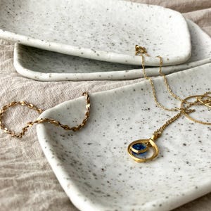 May include: A gold necklace with a blue pendant and a gold chain bracelet rest on a speckled white ceramic tray. The jewelry is displayed on a neutral-colored linen surface, creating a minimalist aesthetic.
