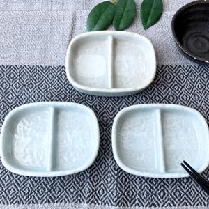 Może przedstawiać: Trzy jasnoniebieskie ceramiczne podzielone talerze. Każdy talerz ma owalny kształt z centralnym dzielnikiem, tworzącym dwie przegródki. Naczynia ułożone są na wzorzystej powierzchni, z czarną miską i pałeczkami w tle.