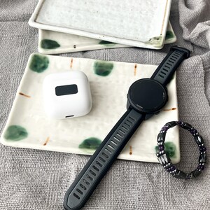 Puede incluir: Un reloj inteligente Garmin negro con una correa negra, una funda blanca para auriculares inalámbricos y una pulsera de cuentas con cuentas moradas y negras se exhiben en un plato decorativo. El plato tiene un fondo blanco con detalles verdes y marrones.
