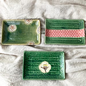 May include: Three rectangular green ceramic plates with floral and geometric designs. One plate features a white flower with yellow stamens, another has a red and white geometric band, and the third has floral patterns.
