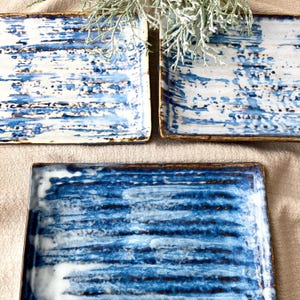 Puede incluir: Tres fuentes rectangulares de cerámica con un diseño abstracto azul y blanco. Las fuentes tienen un borde marrón y una superficie texturizada. El diseño se asemeja a pinceladas. Las fuentes están dispuestas sobre una superficie de color neutro.