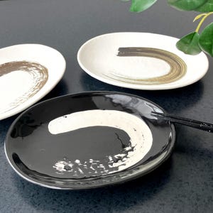 May include: Three ceramic plates with a minimalist design. One plate is black with a white brushstroke, and two are white with brown brushstrokes. Chopsticks rest on the black plate. The plates are oval-shaped.