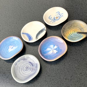 May include: A collection of small, oval-shaped ceramic dishes with hand-painted designs. The dishes feature various patterns, including a crab, a bunny, a flower, and abstract brushstrokes. The color palette includes blue, white, and earth tones.