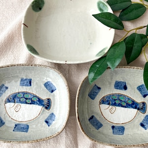 May include: A collection of ceramic plates with a hand-painted design. Two rectangular plates feature a blue and white pufferfish illustration, surrounded by blue squares. A round plate with green dots is also included.
