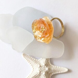 Puede incluir: Un anillo dorado con un diseño de flor grande, translúcida, de color naranja-amarillo y pequeños detalles dorados. El anillo descansa sobre vidrio de mar blanco y una estrella de mar blanca.
