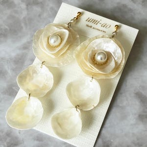 Puede incluir: Pendientes colgantes con pétalos de concha superpuestos en color blanco roto translúcido y una perla blanca central con un detalle de alambre dorado. Los pendientes están sujetos a una tarjeta blanca con el texto umi-ao Shell accessories.