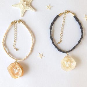May include: Two braided cord necklaces with seashell pendants. One is cream, the other dark grey. Both have gold-coloured clasps and adjustable chains. The pendants are flower-shaped with pearl accents. Starfish and small star-shaped decorations are scattered around the necklaces.