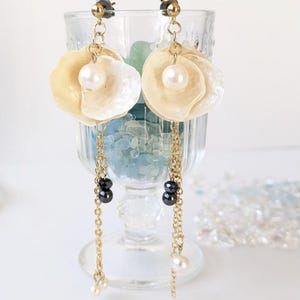 Op de afbeelding: Elegante oorhangers met crèmekleurige, bloemvormige schelpen met een parel in het midden. Gouden kettingen hangen zwarte kralen en kleine parels op. De oorbellen worden in een helder glas getoond.