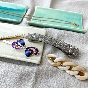 Op de afbeelding: Een assortiment haaraccessoires en sieraden, waaronder een gouden ketting, een parel haarclip, een paarse en blauwe juwelen haarclip, een met kristallen bezette haarspeld en een haarclip met schakels. De items worden gepresenteerd op kleine, rechthoekige keramische trays.