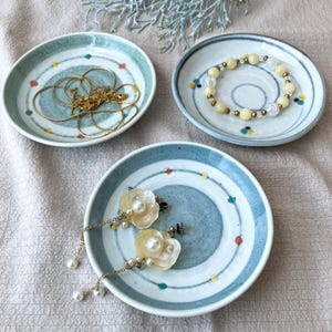 May include: Three small, round ceramic dishes with a blue and white spiral design. One dish holds a gold necklace, another a beaded bracelet, and the third, a pair of flower-shaped earrings with pearl accents. The dishes are on a neutral-colored surface.
