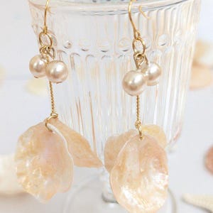 May include: Gold-coloured drop earrings with two faux pearls and a fine chain, suspending a pearlescent, petal-shaped charm. The earrings have a light peach and cream colour scheme, evoking a natural, elegant aesthetic. These earrings are a perfect accessory for any occasion.