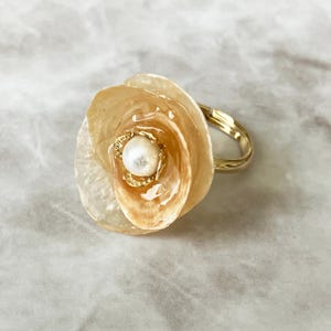 May include: A gold-toned ring featuring a large, translucent, beige-colored flower-shaped ornament. The center of the flower is adorned with a small, round, white pearl set within a delicate gold-colored metal frame with a smooth, polished finish.