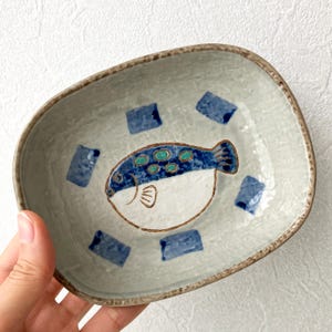 May include: A ceramic, rectangular dish with a hand-painted fish design. The fish is white with blue and teal accents, and the dish has a light gray background with blue square accents. The rim is a dark brown color.