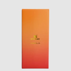 May include: A rectangular product box featuring a vertical gradient that transitions from a bright orange at the top to a deep red at the bottom. The center of the box displays the gold-colored Stanley logo and the text KAROL G printed below it.