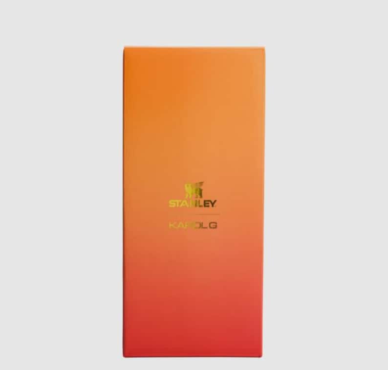 May include: A rectangular product box featuring a vertical gradient that transitions from a bright orange at the top to a deep red at the bottom. The center of the box displays the gold-colored Stanley logo and the text KAROL G printed below it.