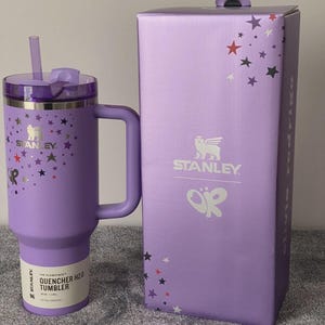 May include: A light purple Stanley Quencher H2.0 tumbler with a matching handle, straw, and lid. The tumbler features a star pattern and the text 'OR' with a butterfly logo. It is accompanied by a matching purple box with the same star and logo design.