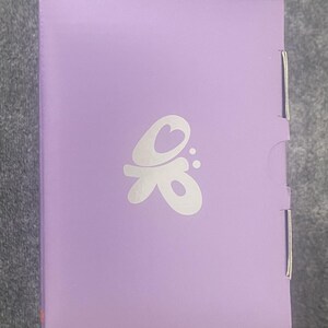 May include: A rectangular lavender-colored box featuring a white stylized graphic logo centered on the front panel. The logo consists of a heart shape integrated into a flowing, abstract design with two small dots to the right. The box has a smooth finish.