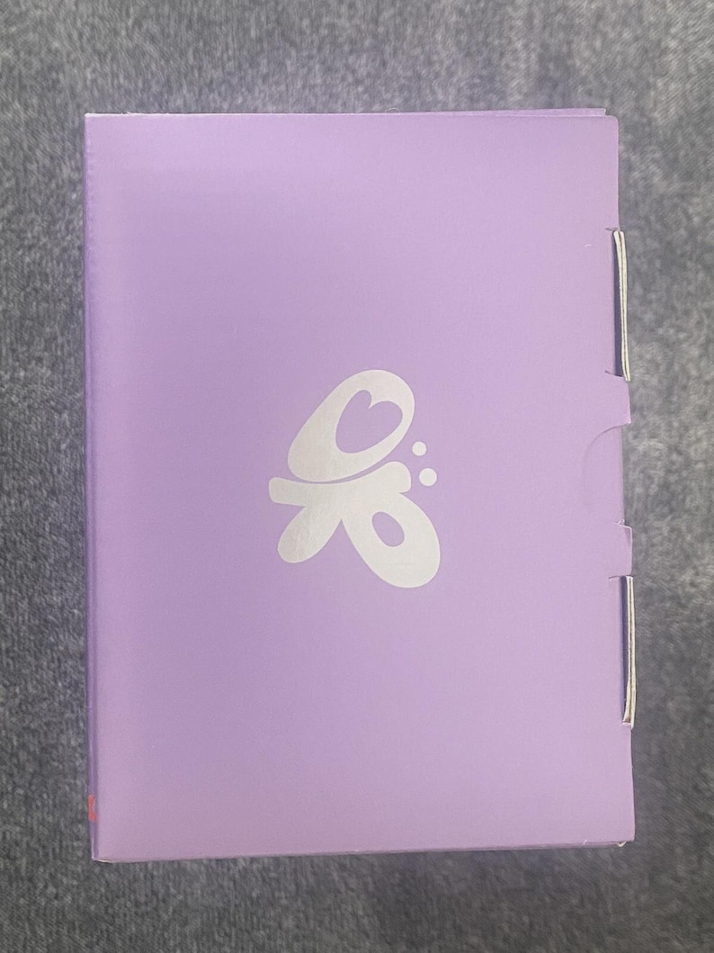 May include: A rectangular lavender-colored box featuring a white stylized graphic logo centered on the front panel. The logo consists of a heart shape integrated into a flowing, abstract design with two small dots to the right. The box has a smooth finish.