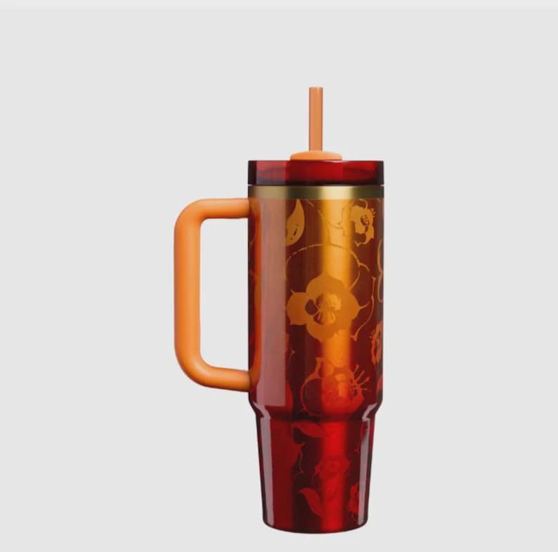 May include: A tall, cylindrical tumbler with a deep red and orange gradient finish featuring a subtle floral pattern. It includes a large, thick orange handle on the side, a matching orange straw protruding from the top, and a gold-toned metal rim.